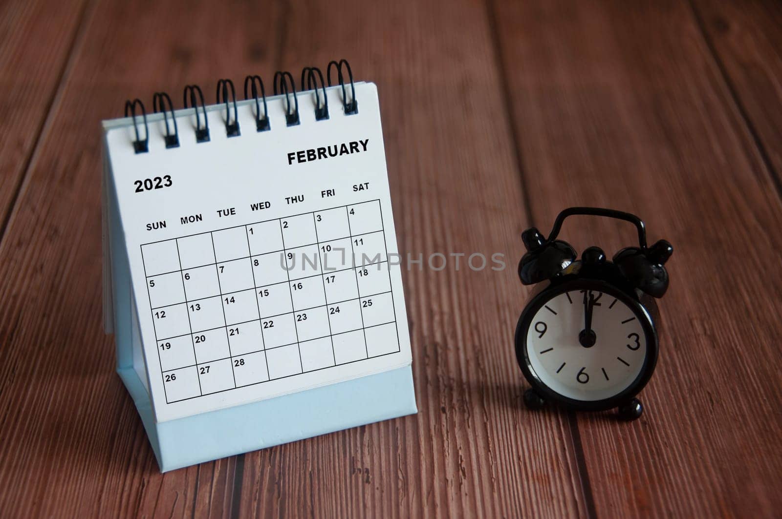 February 2023 white desk calendar on wooden table with alarm clock pointing at 12 o'clock. Calendar concept