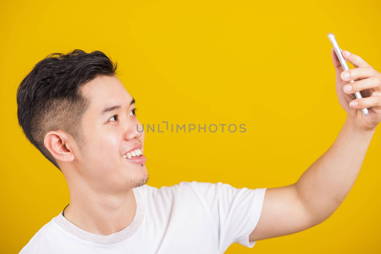 Asian handsome young man smiling positive saying hi talking by video call with mobile phone, excited happy male taking selfie on smartphone studio shot isolated on yellow background