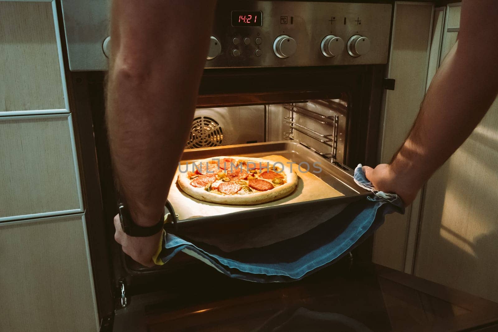 A man bakes pizza at home using an oven. Men's hands pull a pizza baking sheet out of the oven
