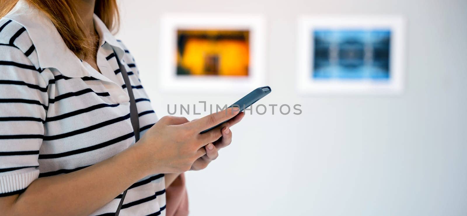 Asian woman standing takes picture art gallery collection in front framed paintings pictures on white wall with mobile phone, people watch at photo frame with smartphone at artwork gallery show