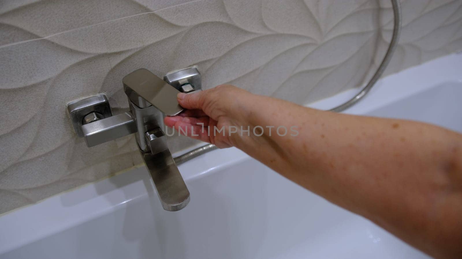 old woman hand is going to turn off the faucet water valwe in the bathroom with biege interior.