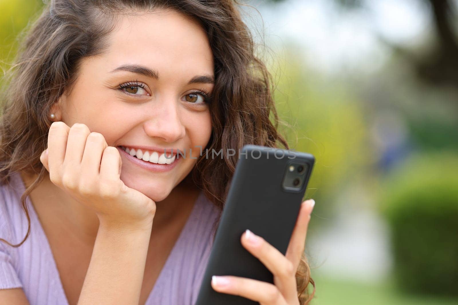 Happy beautiful woman holding cell phone looks at you in a park
