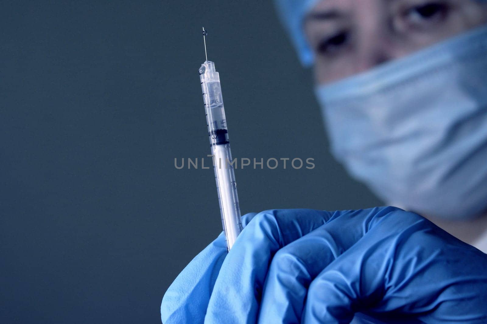 A woman doctor in a medical cap and mask holds a medical syringe in her hand. Soft focus..