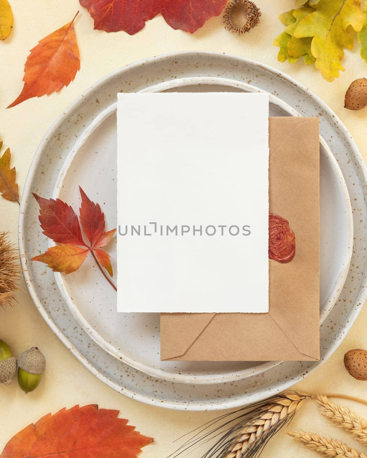 Autumn table place with colorful leaves and blank card with envelope top view. Mockup of fall vertical card on light yellowtable, flat lay, copy space. Harvest, Halloween and Thanksgiving season