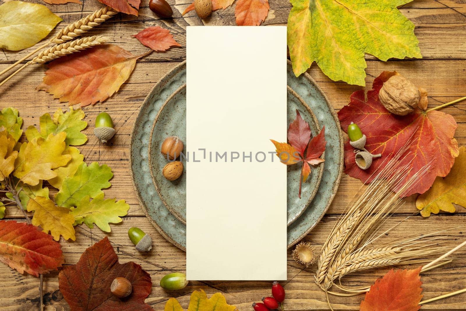 Autumn rustic table setting with blank card between colorful leaves and berries, top view. Vertical menu card mockup on wooden table, copy space. Thanksgiving, halloween or harvest concept