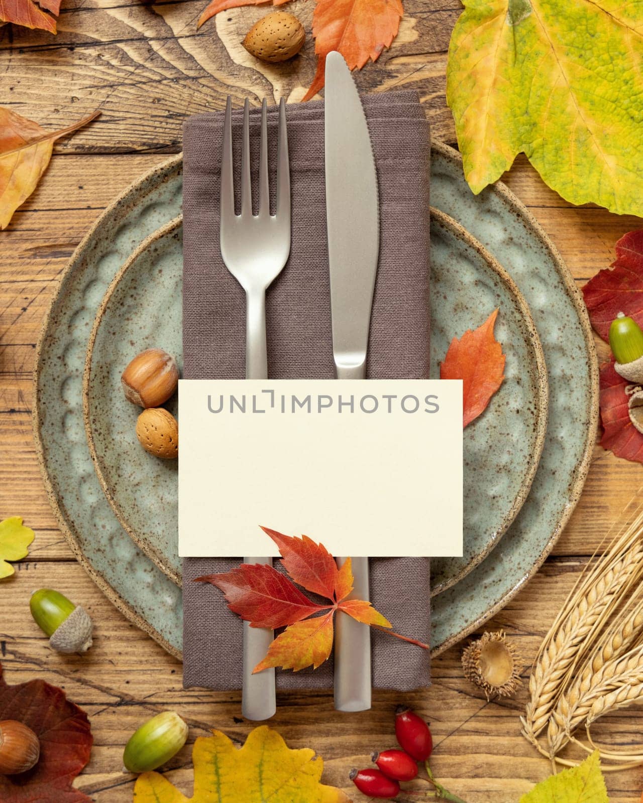 Autumn rustic table place with blank card between colorful leaves and berries, top view, mockup. Flat lay with horizontal card on wooden table, copy space. Thanksgiving or harvest concept