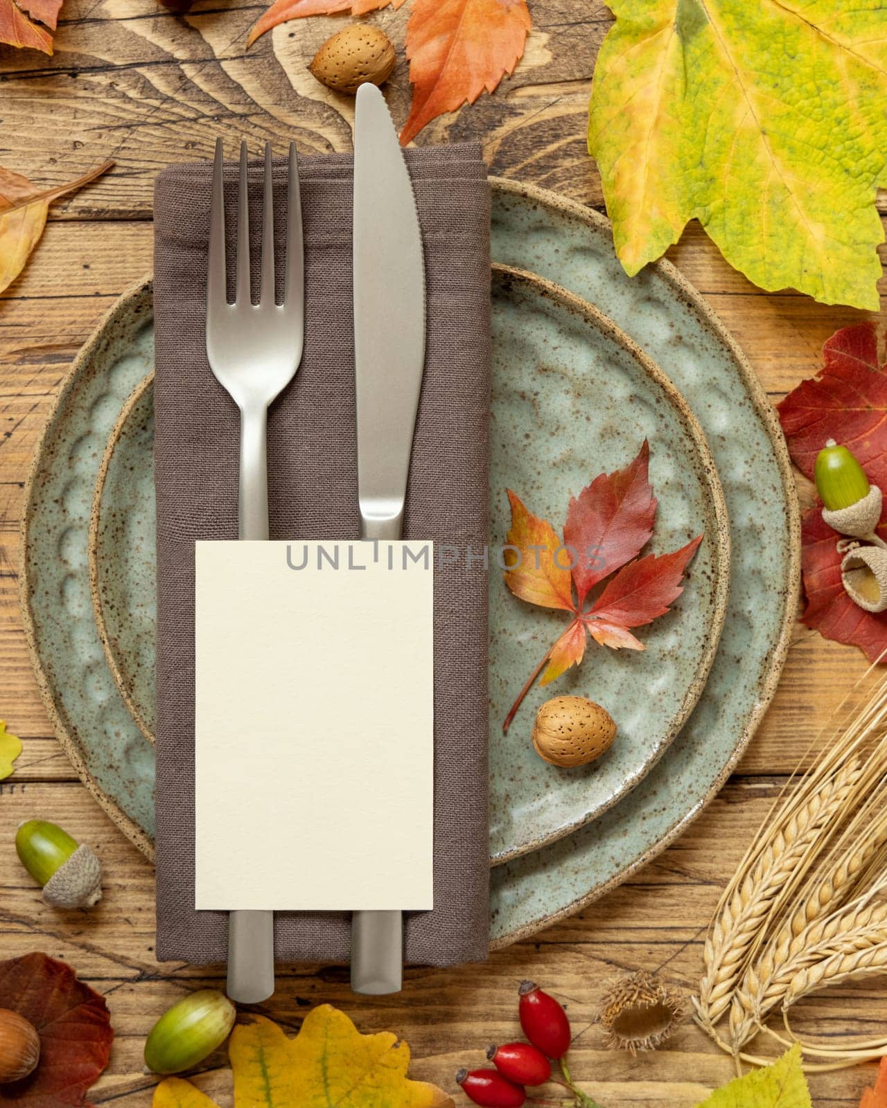 Autumn rustic table place with blank card between colorful leaves and berries, top view, mockup. Flat lay with vertical card on wooden table, copy space. Thanksgiving or harvest concept