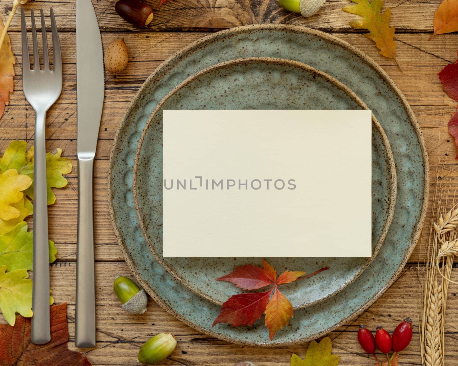 Autumn rustic table setting with blank card between colorful leaves and berries, top view. Horizontal card mockup on wooden table, copy space. Thanksgiving, halloween or harvest concept