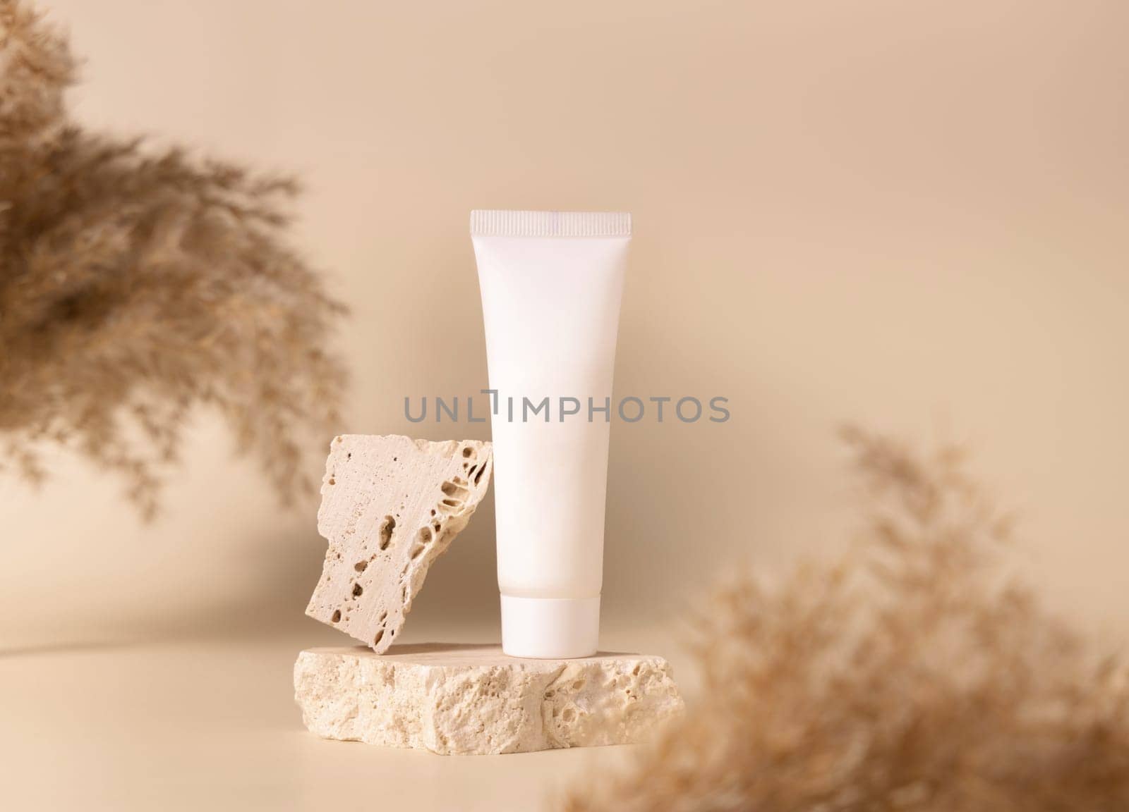 White plastic cream tube on travertine stone near dry pampas grass on light yellow, close up, mockup. Skincare beauty product. Natural cosmetics. Boho composition..
