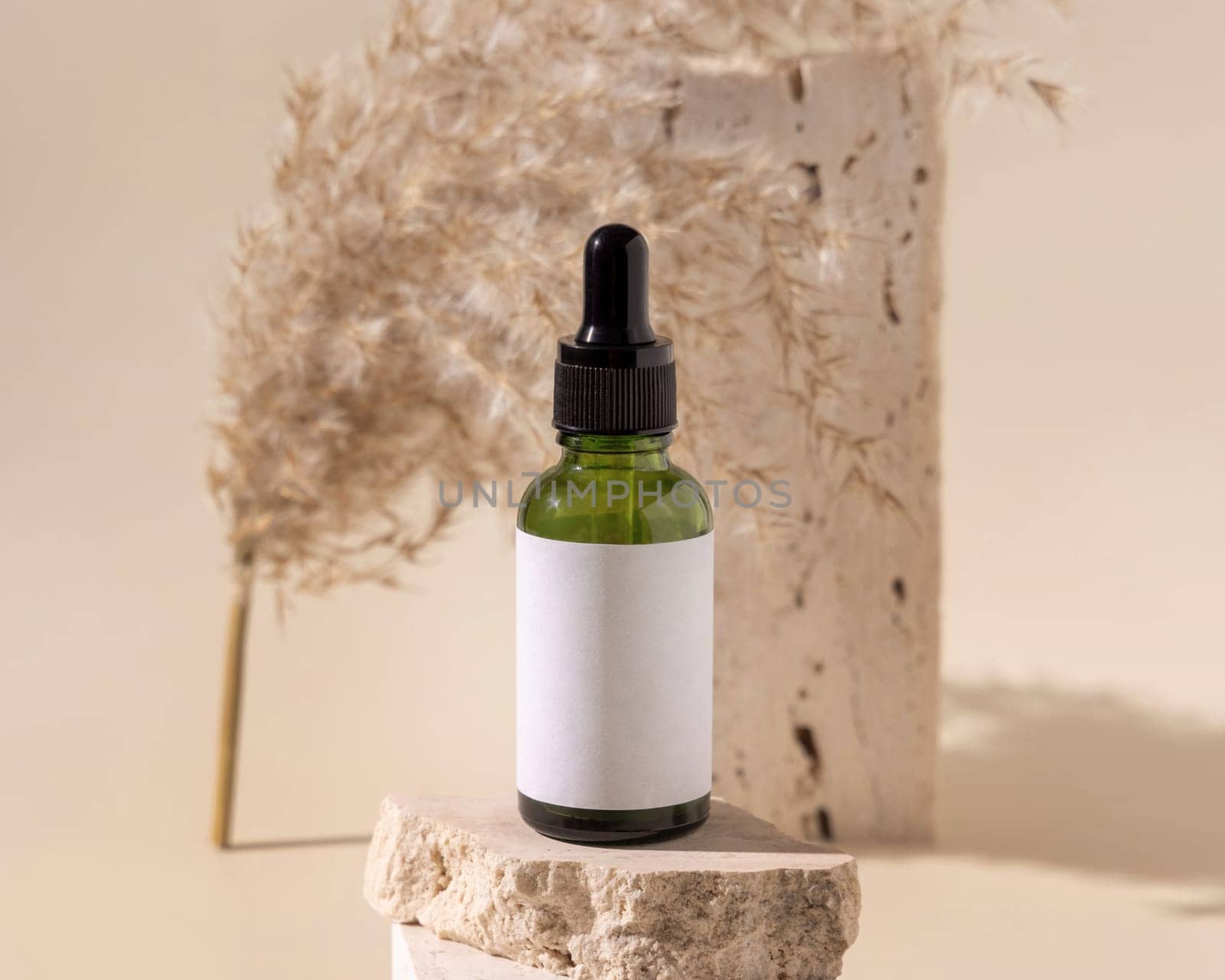 Green dropper bottle on travertine stone near dry pampas grass on light yellow, close up, mockup. Skincare beauty product. Natural cosmetics. Boho composition..