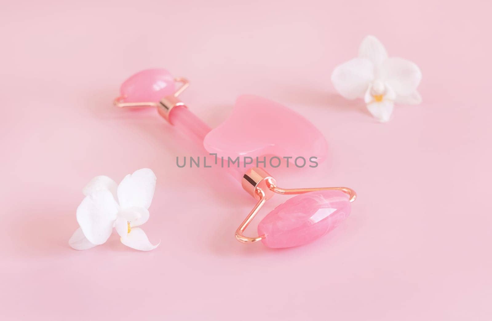 Rose quartz facial roller and gua sha massager near white orchid flowers, close up on light pink. Anti-age massage and natural lifting therapy at home. Facial skin care at home. Beauty concept