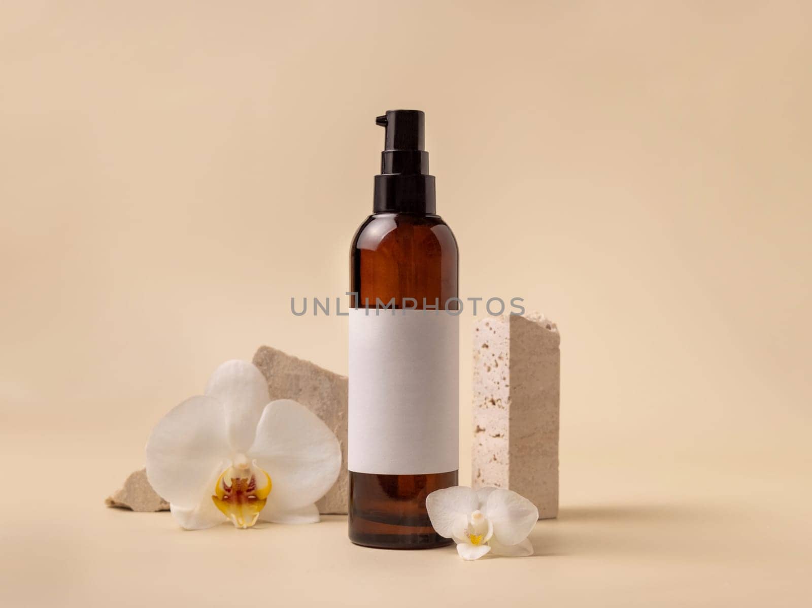 Dark glass cosmetic bottle with blank label near white orchid flowers and stones on light yellow, close up, mockup. Skincare handmade beauty product, tonic or lotion. Exotic natural cosmetics..