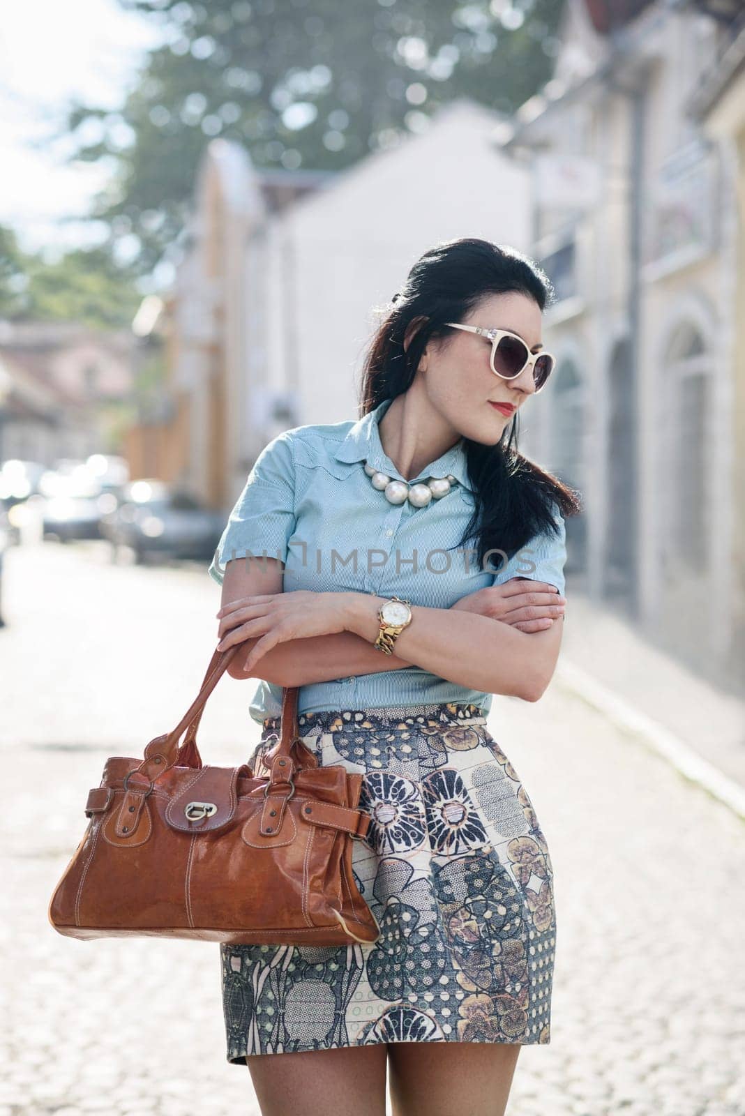 Beautiful stylish woman in elegant smart fashion clothes posing on the street. Wearing fashionable coclourful skirt, blue shirt, sunglasses, pearl neckles and holding leather handbag.