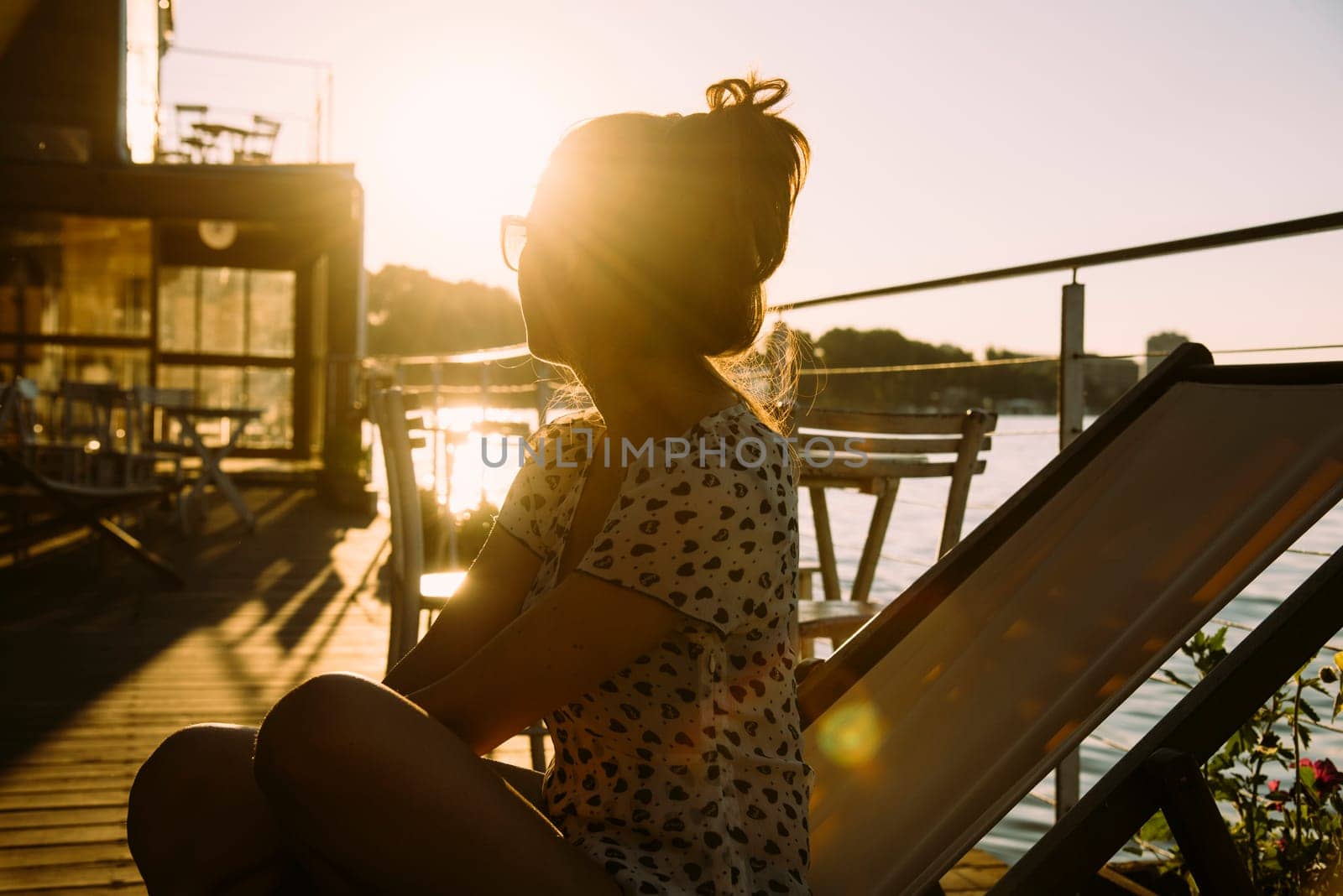 Young hipster woman in summer clothing sitting in cafe, enjoys the sun and watching the river at summer sunset. Retro style image.