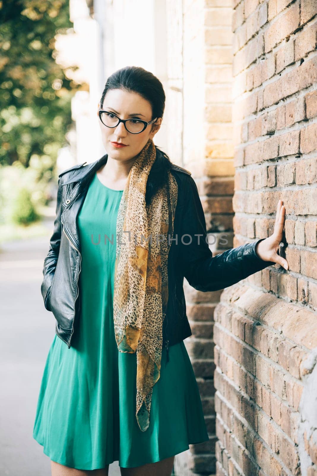 Fashionable young woman in leather jacket and glasses behind brick wall on the street.
