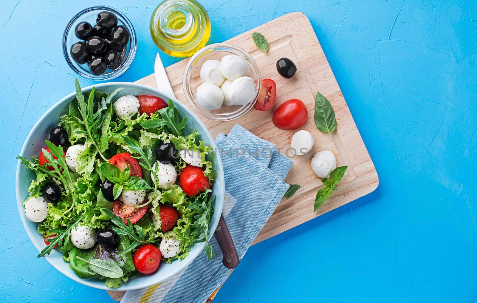 Preparation of Green Salad with mozzarella cheese, black olives, arugula and tomato on blue table background