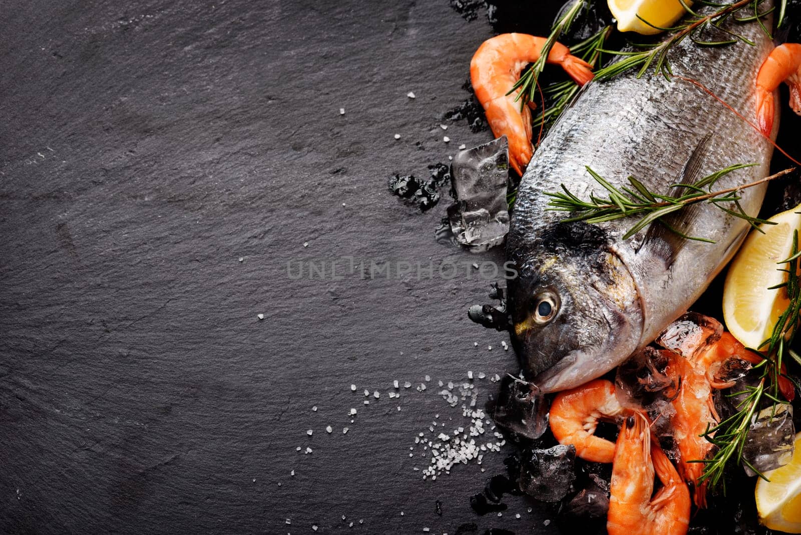 Top view of fresh sea food on grunge textured black background. Shrimps, bream, herbs, lemon and ice on slate with copyspace.