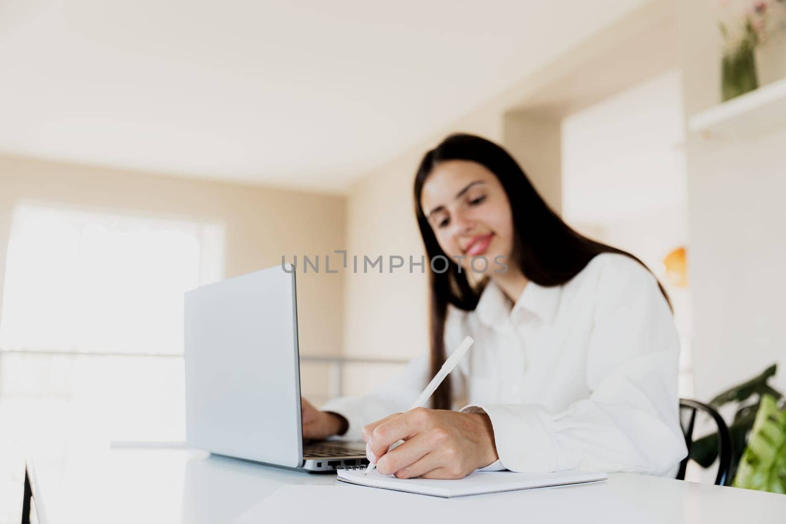 Caucasian business woman sitting at desk, reading from computer, taking notes at notebook. Female student using pc, writing down important information with pencil in note pad. Happy smiling lady