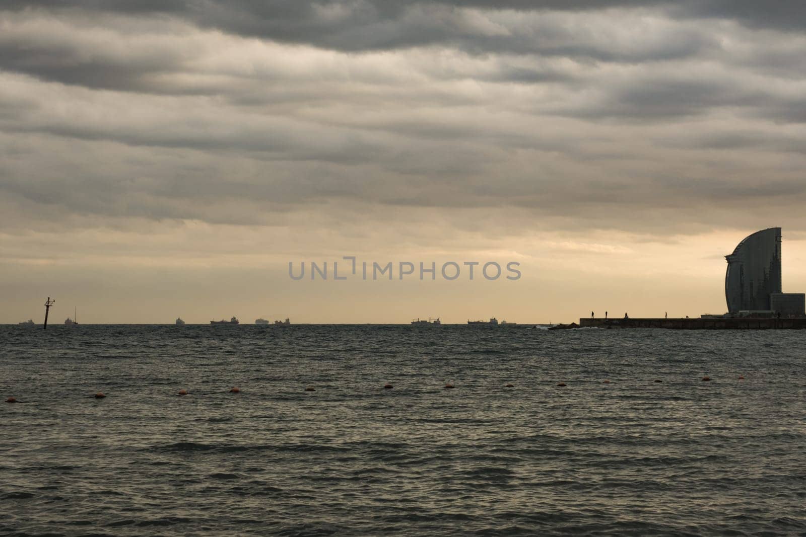 Movement of cargo ships near the port of Barcelona and silhouettes of some people walking on the quay to enjoy a suggestive autumn twilight.