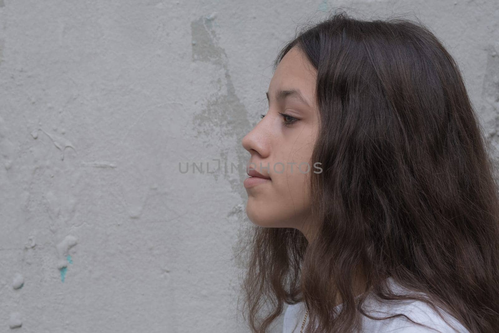 Close up profile of a teenage girl with long brown hair looks forward with a thoughtful expression. Background of gray wall, copy free space for text.