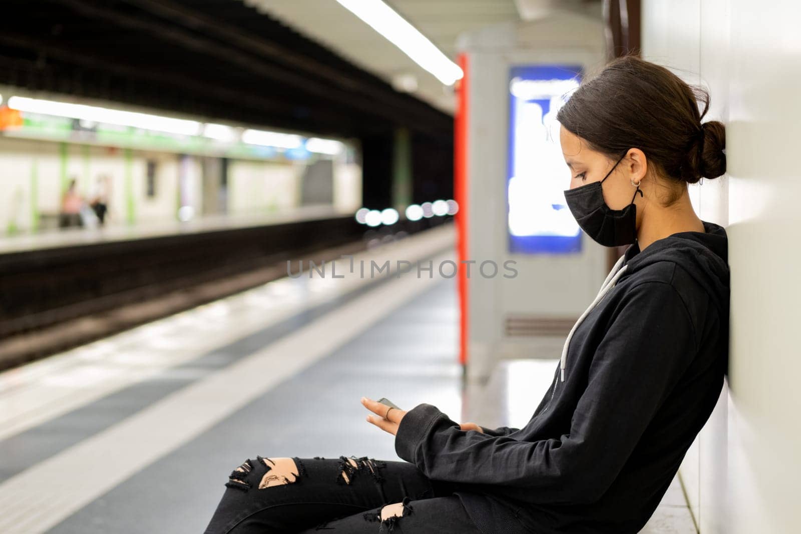 Teenage girl sitting on a bench at subway station. She's wearing a sweatshirt and black ripped jeans. Some people who use public transport still use protective masks that completely cover their mouth and nose.
