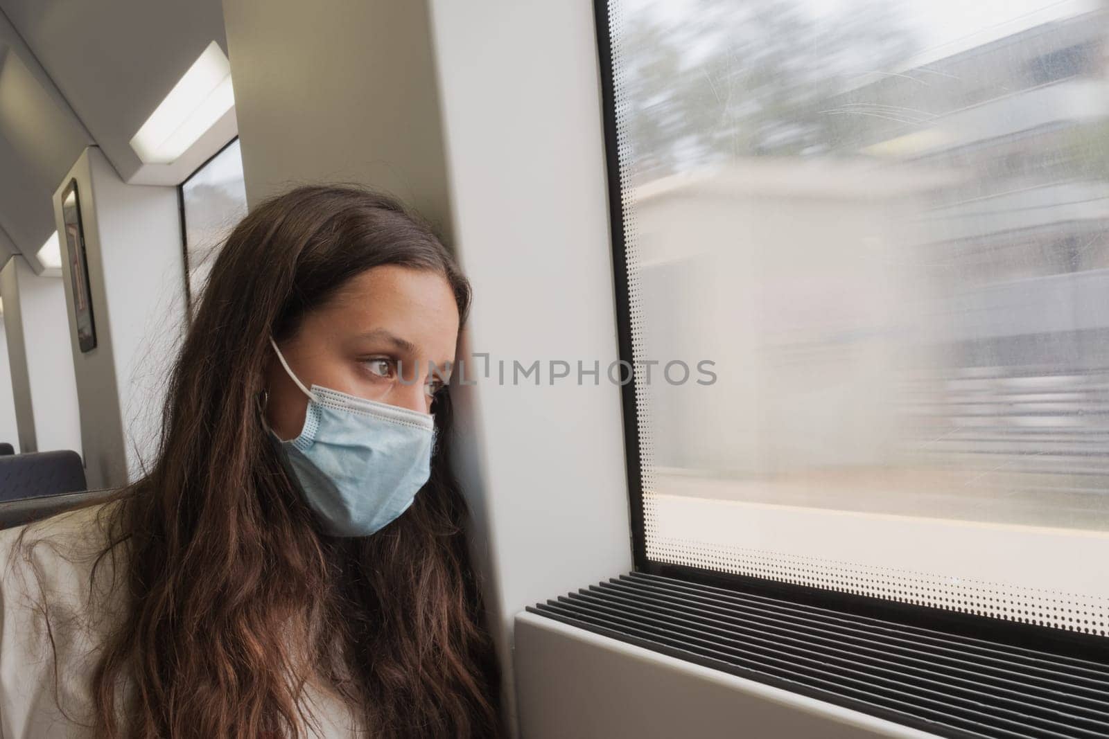 Pretty girl with long hair sitting in a local train looking outside the window. A young train passenger with protective mask. She seems to be traveling with her mind as well and her green eyes are looking very far away.