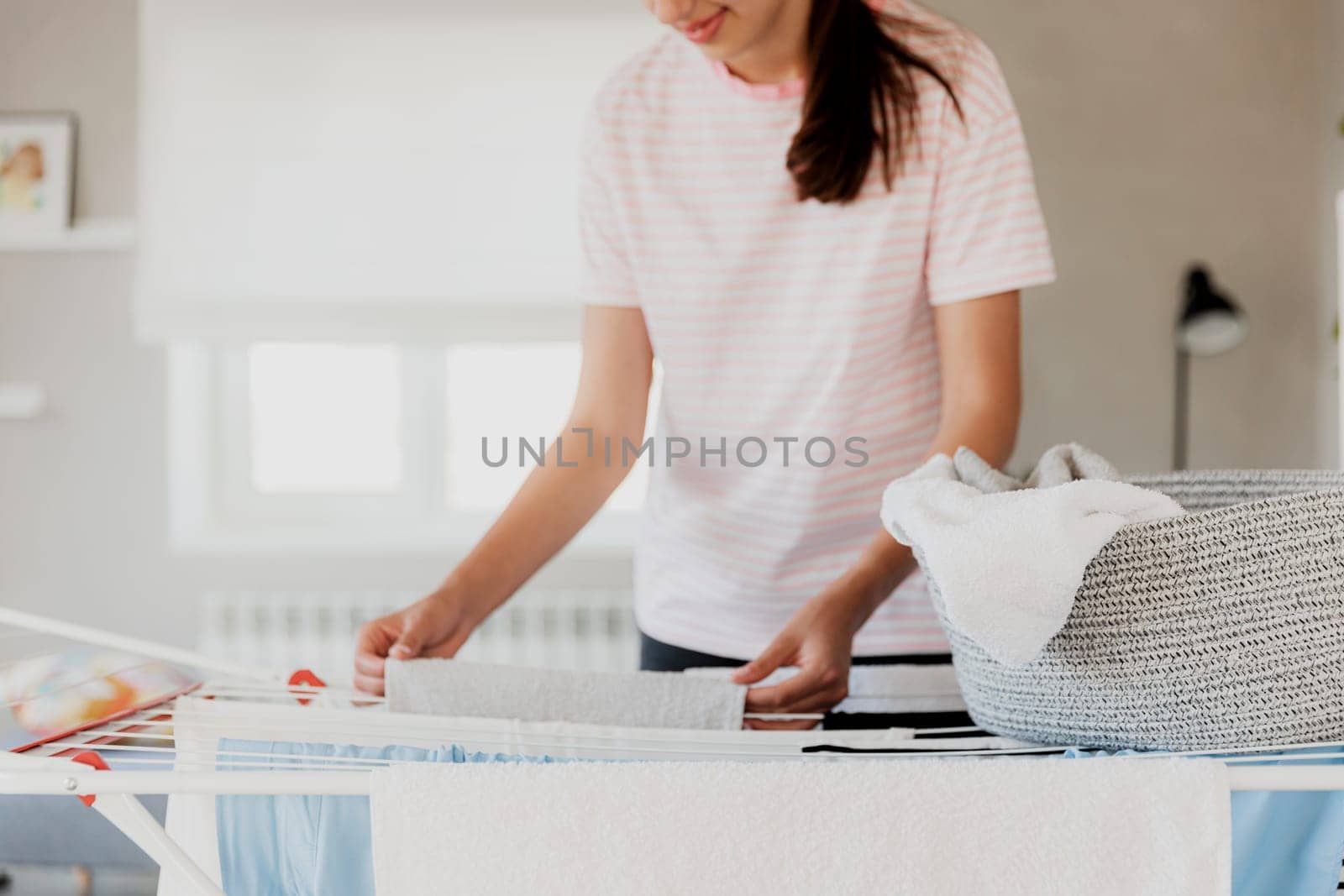 Happy young brunette caucasian woman smiling, hanging clean wet clothes laundry on drying rack at home. Female housewife hands closeup spreading laundry from basket. Girl puts towels after drying them