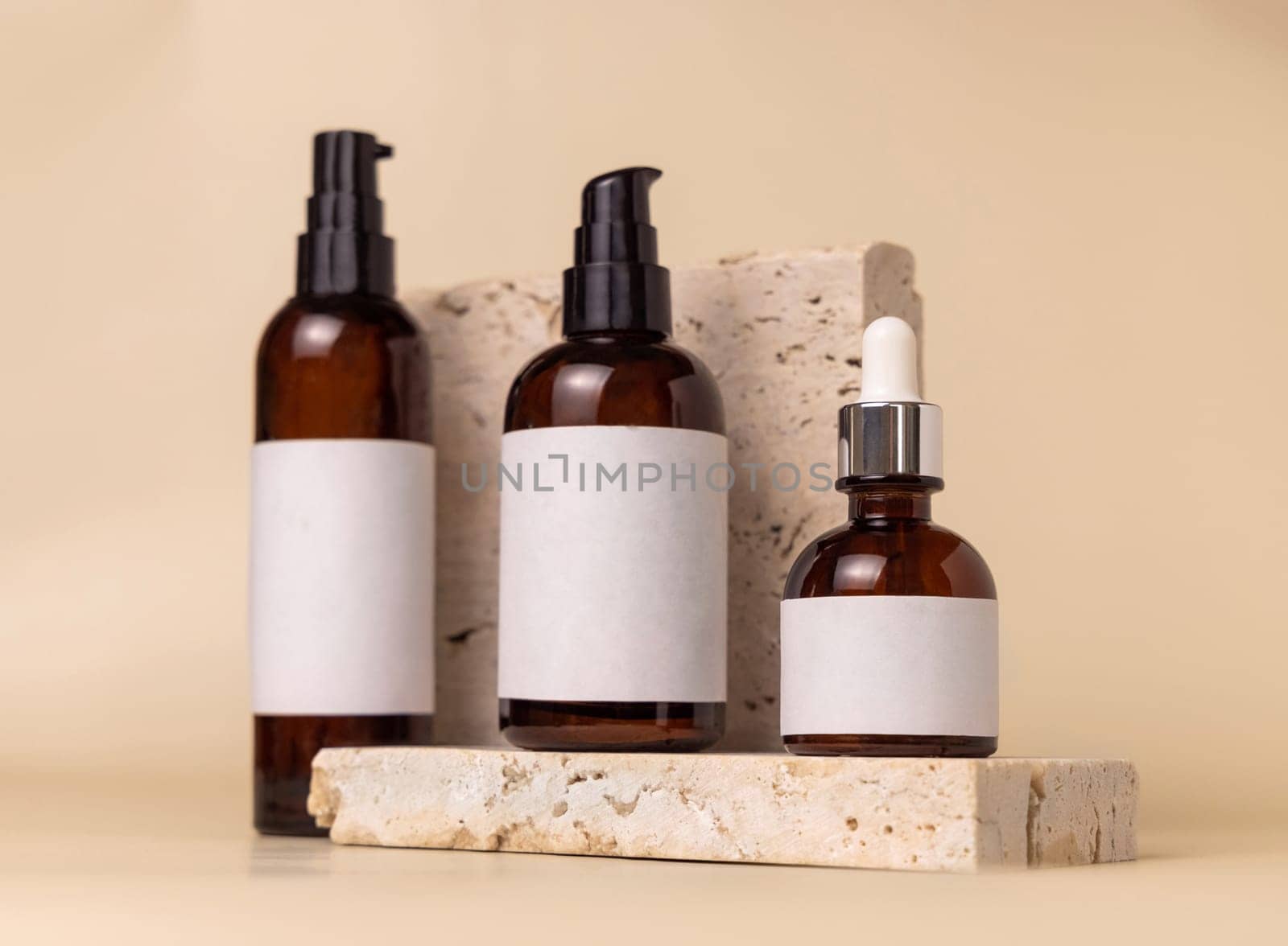 Brown glass one pump bottles on travertine stones on light beige close up, label mockup. Skincare beauty product package. Natural cosmetics, minimal composition..