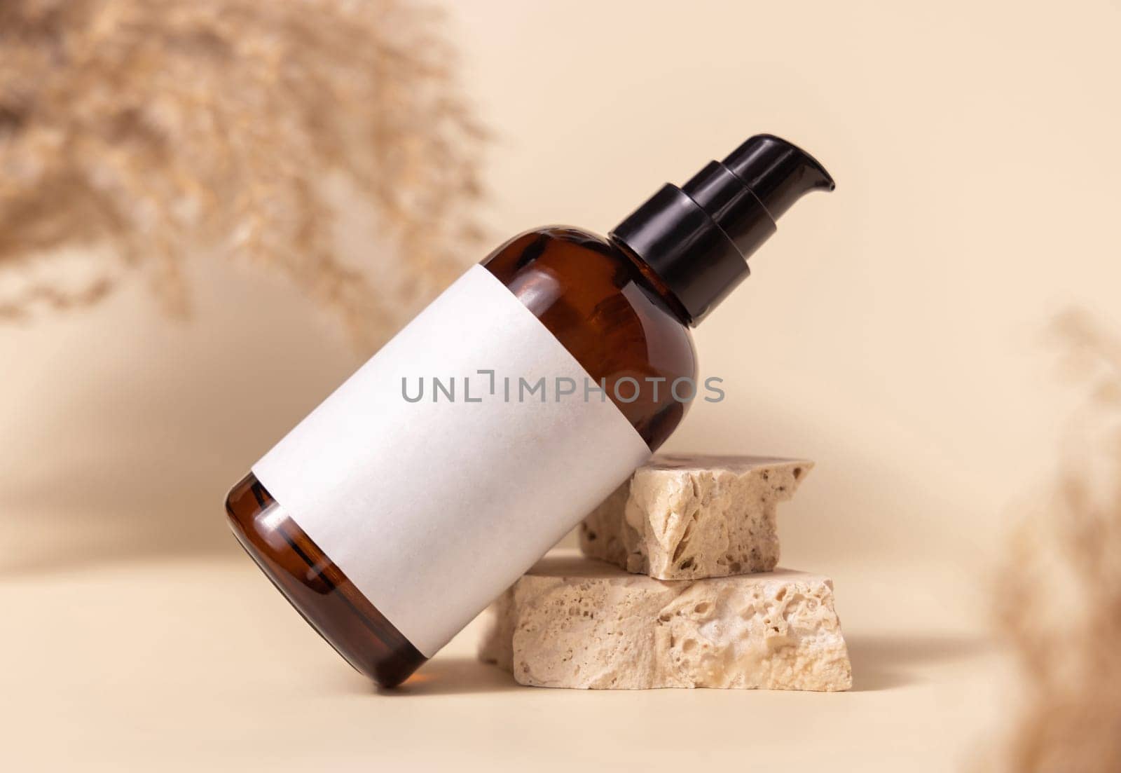 Brown glass cosmetic bottle with blank label on stones near pampas grass on light yellow, close up, mockup. Skincare beauty product. Natural eco friendly cosmetics