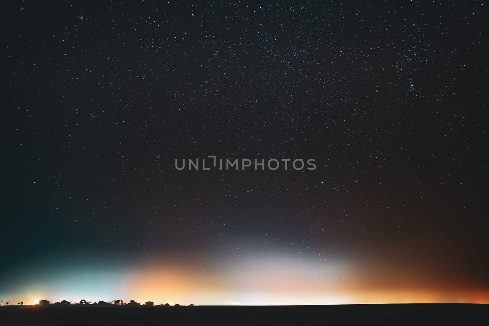 Night Starry Sky With Glowing Stars Above Countryside Town Landscape. Rural Field Meadow In Early Spring. Bright Glow Of Stars. Illumination Lights Of Small Town Or Village.