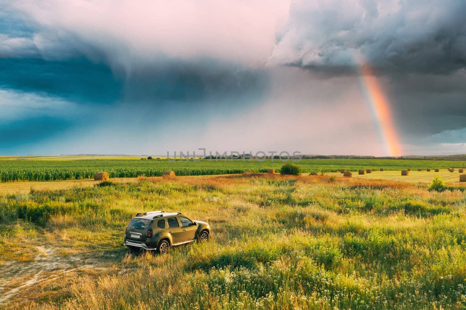 Elevated View Of Car SUV Parked Near Countryside Road In Summer Field Rural Landscape. Altered Rainy Sky With Rinbow Above Meadow Rural Landscape.