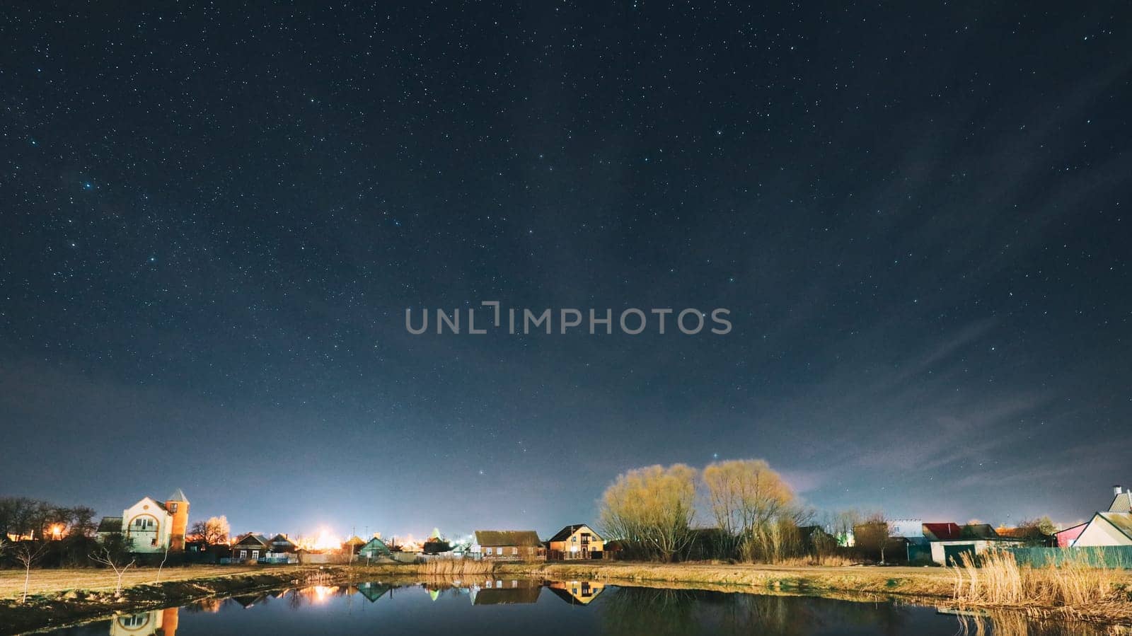 Belarus, Eastern Europe. Night Sky Stars Above Countryside Landscape With Lake Coast And Small Town Or Village. Natural Starry Sky Above Pond And Houses In Early Spring Night. Panorama Panoramic View