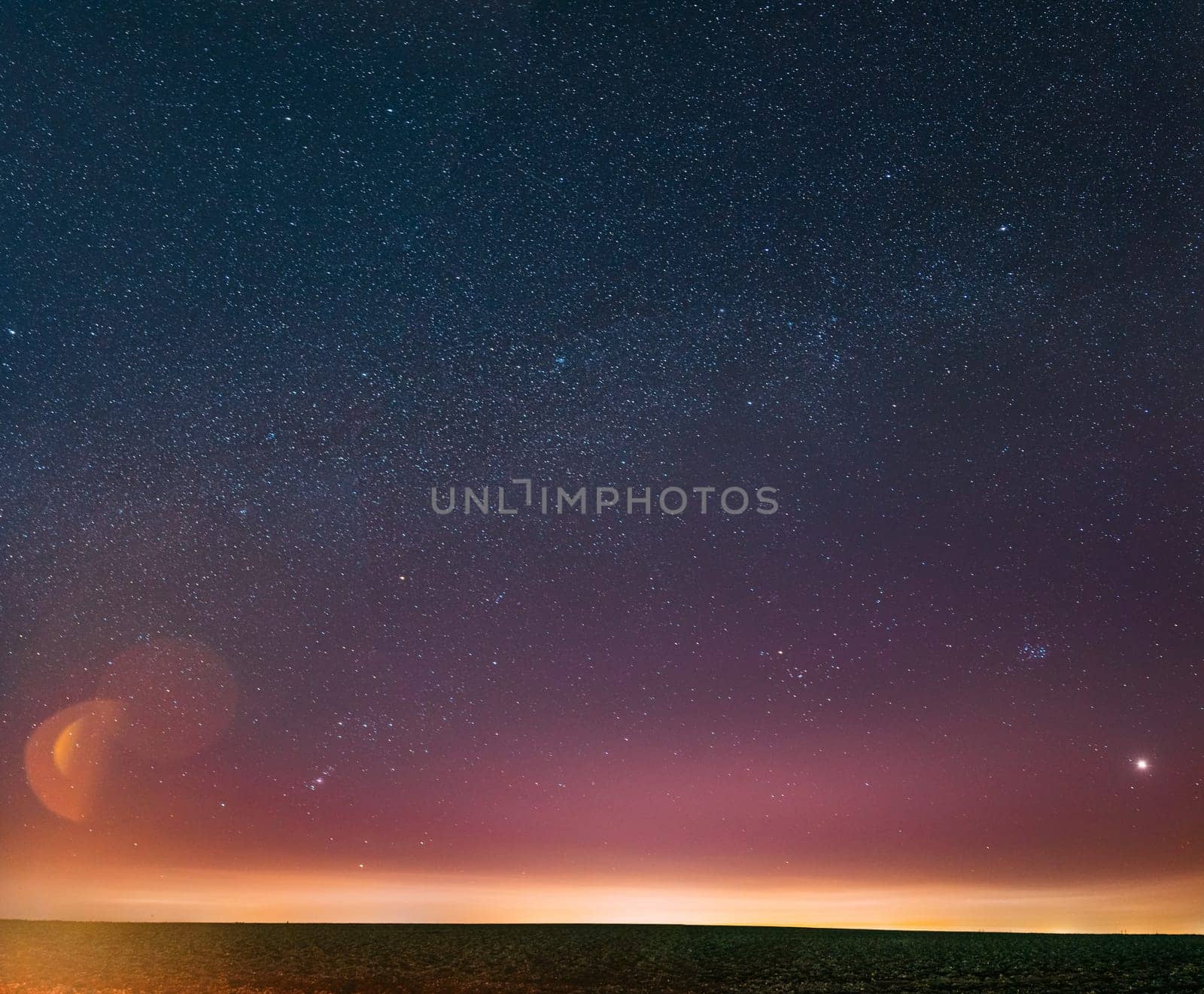 Night Starry Sky With Glowing Stars Above Countryside Landscape. Milky Way Galaxy And Rural Field Meadow In Early Spring. Bright Glow Of Planet Venus Among The Stars. Warm Lights Of Evening Sunset.