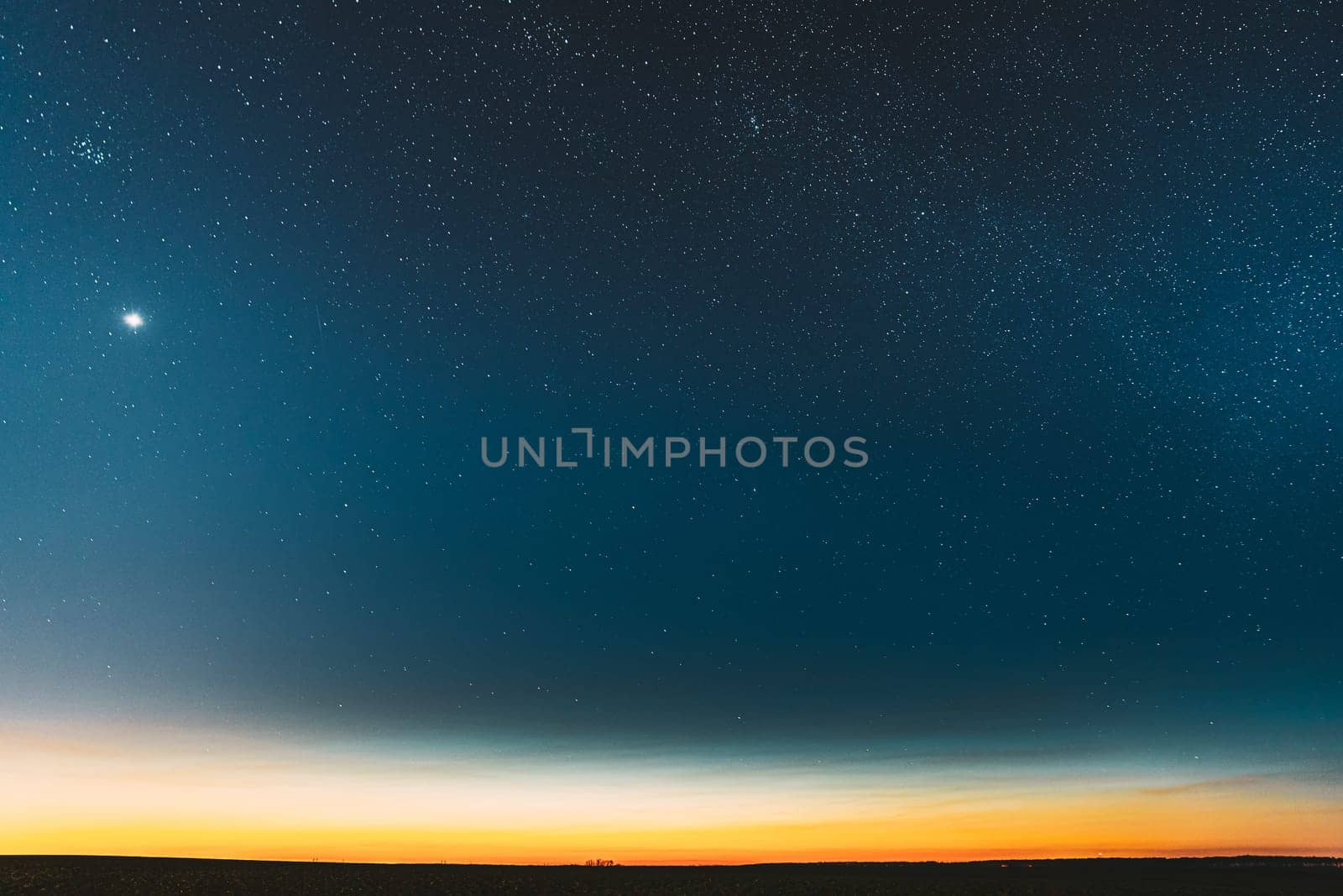 Night Starry Sky With Glowing Stars Above Countryside Field Landscape In Early Spring. Bright Glow Of Planet Venus In Sky Among The Stars. Sky In Warm Lights Of Evening Sunset Dawn Or Morning Sunrise.
