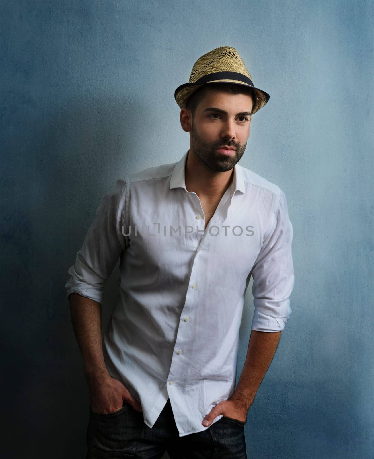 Young sexy hipster guy with beard posing in front of blue grunge wall. Portrait of stylish male model with retro hat and casual style clothing. Man's fashion and beauty.