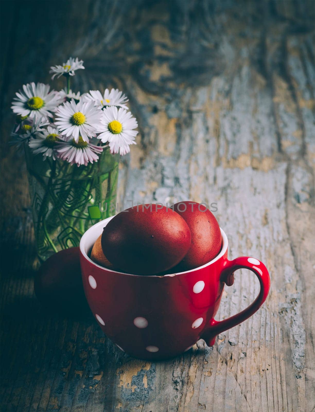 Easter still life on dark background. Hand painted red easter eggs in retro jug and daisy flower bouquet on rustic wooden table fine art.  Traditional easter holiday celebration concept.