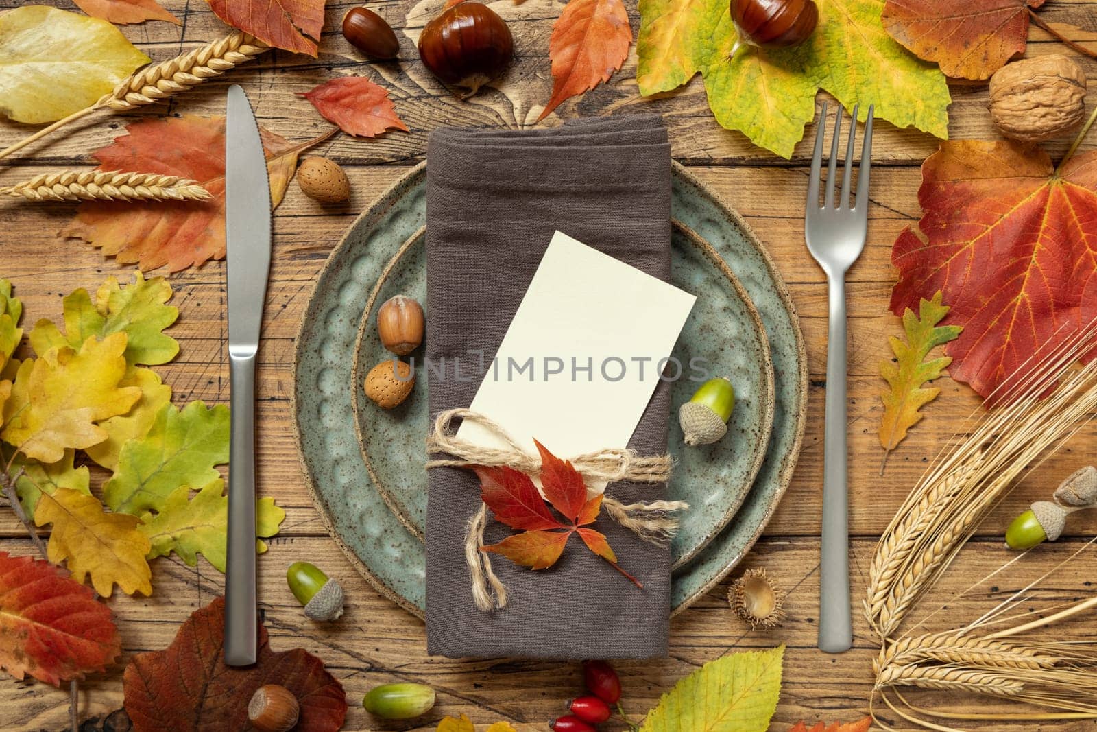 Autumn rustic table place with blank card between colorful leaves and berries, top view, mockup. Flat lay with vertical card on wooden table, copy space. Thanksgiving or harvest concept