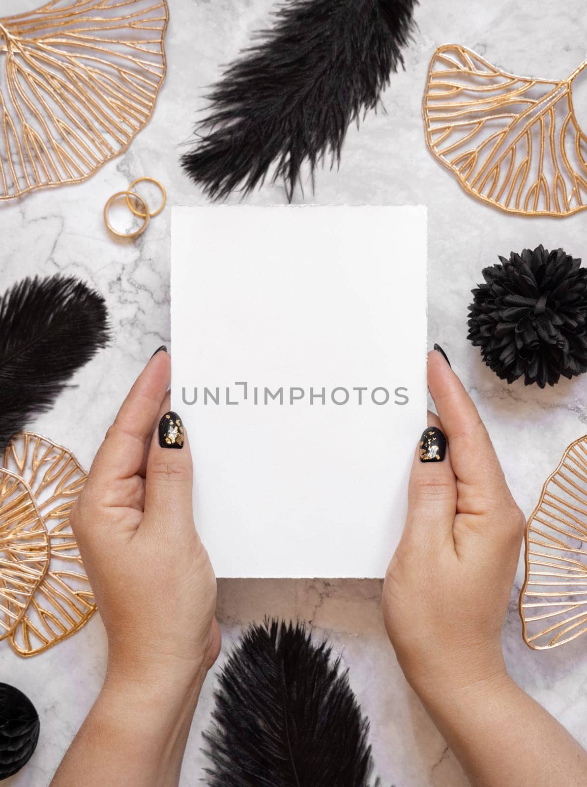 Hands with Blank card near black feathers, golden leaves and wedding rings top view. Invitation or greeting card mockup. Elegant femminine flat lay with vertical paper card