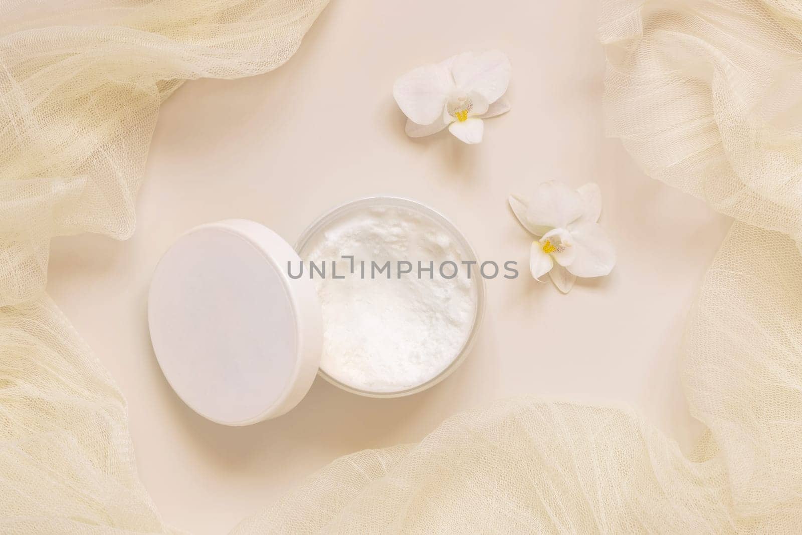 Opened Cream jar with a lid near white orchid flowers and tulle on light yellow top view. Cosmetic Mockup. Exotic natural cosmetics, romantic composition, everyday skincare routine..