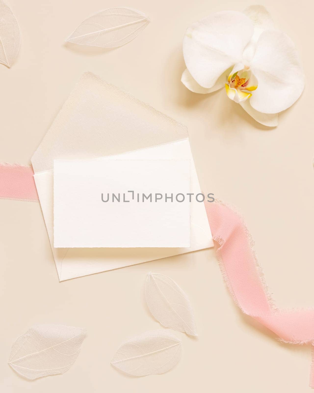 Wedding Card over envelope near white orchid flower and silk ribbons on light yellow top view, mockup. Tropical scene with horizontal blank card flat lay