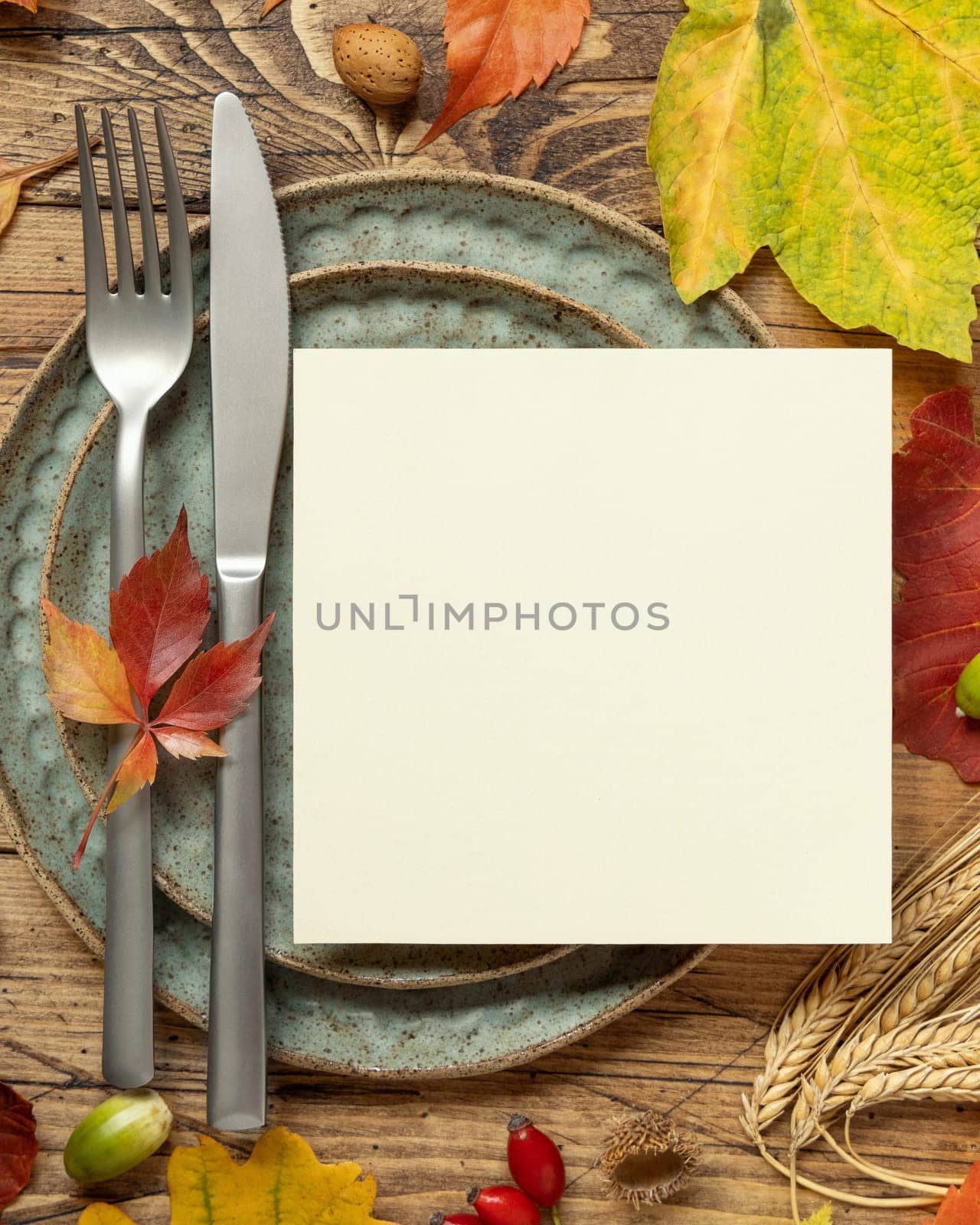 Autumn table setting with red, yellow and orange leaves, berries and blank card top view. Mockup of fall square card on wooden table flat lay copy space. Harvest, Halloween and Thanksgiving season