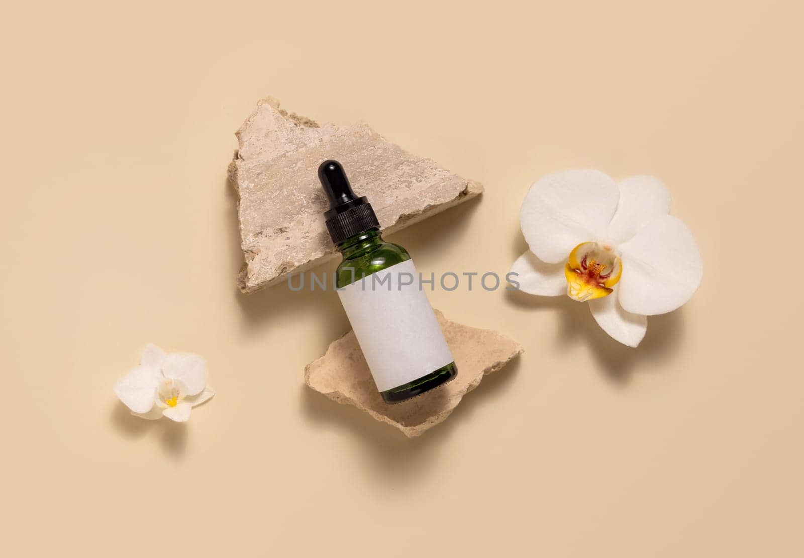 Green glass dropper bottle on stones near white orchid flowers on light yellow, top view, mockup. Skincare beauty product, serum or essential oil. Exotic natural cosmetics..