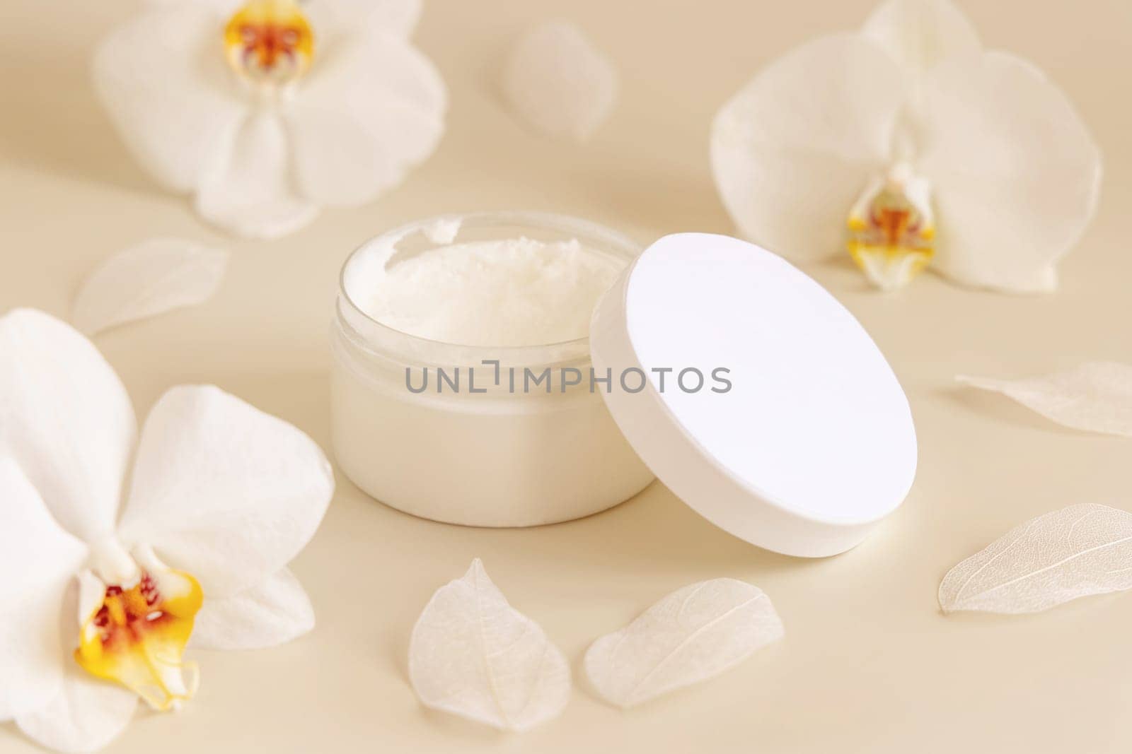 Opened Cream jar with a blank white lid near white orchid flowers on light yellow close up. Cosmetic packaging Mockup. Exotic natural cosmetics, romantic composition, everyday skincare routine..