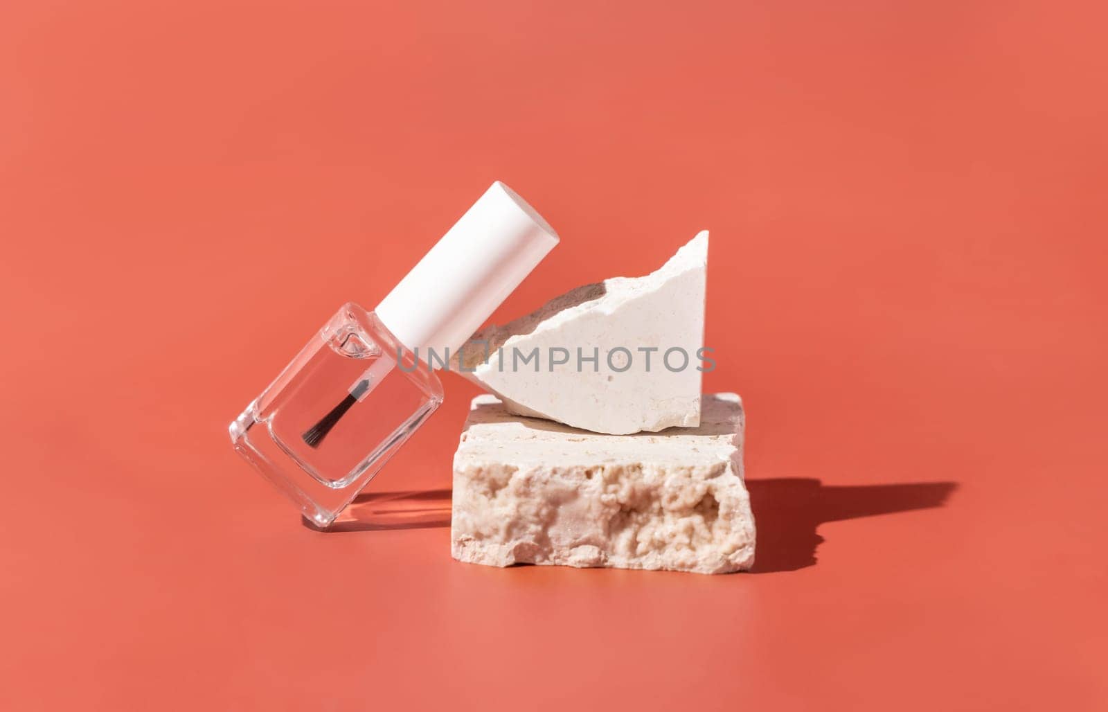 Clear Glass Refillable Bottle with Brush Cap near beige travertine stones on pink, close up, mockup. Skincare nail product. Natural cosmetics, minimal composition..