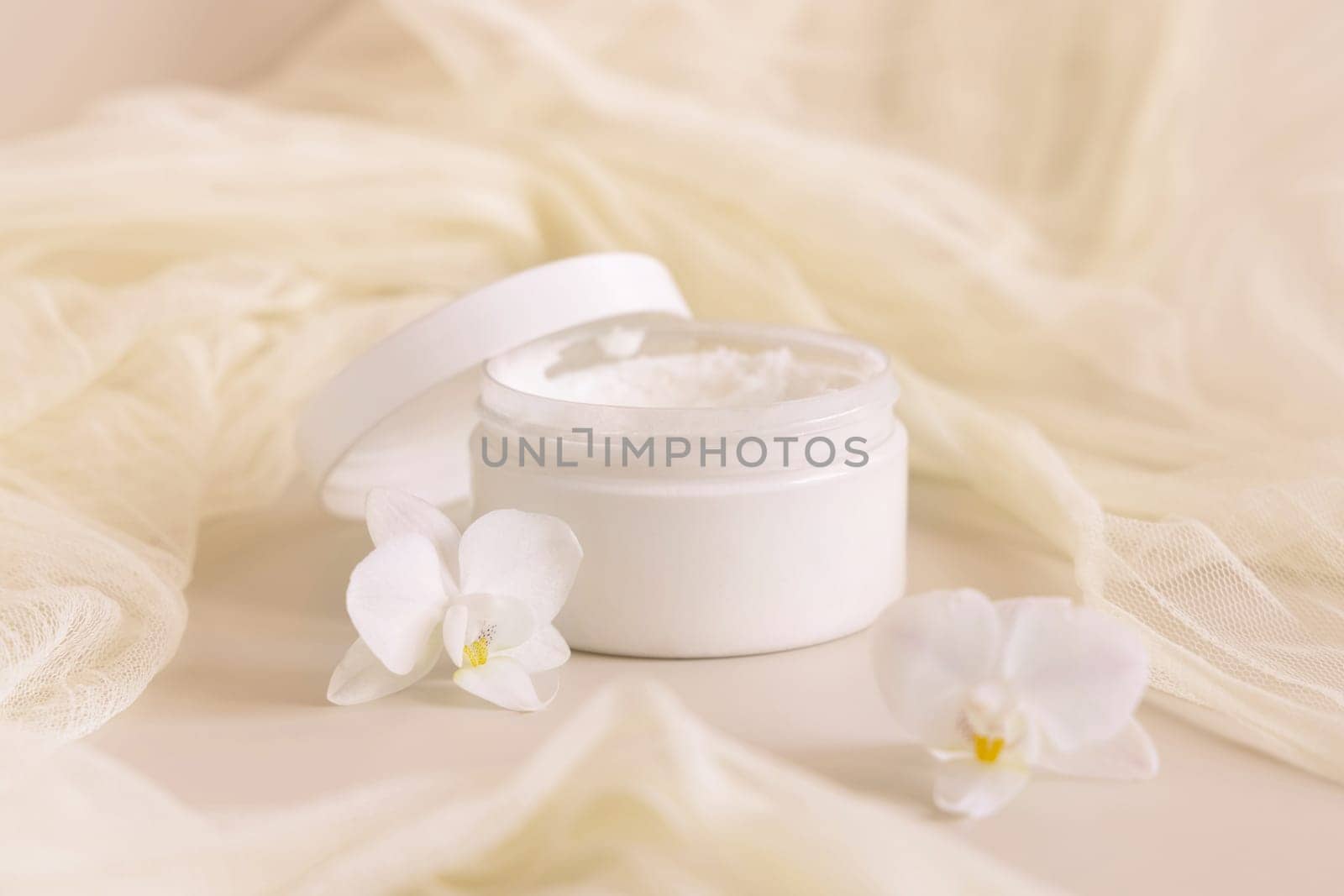 Opened Cream jar with a lid near white orchid flowers and tulle on light yellow top view. Cosmetic Mockup. Exotic natural cosmetics, romantic composition, everyday skincare routine..