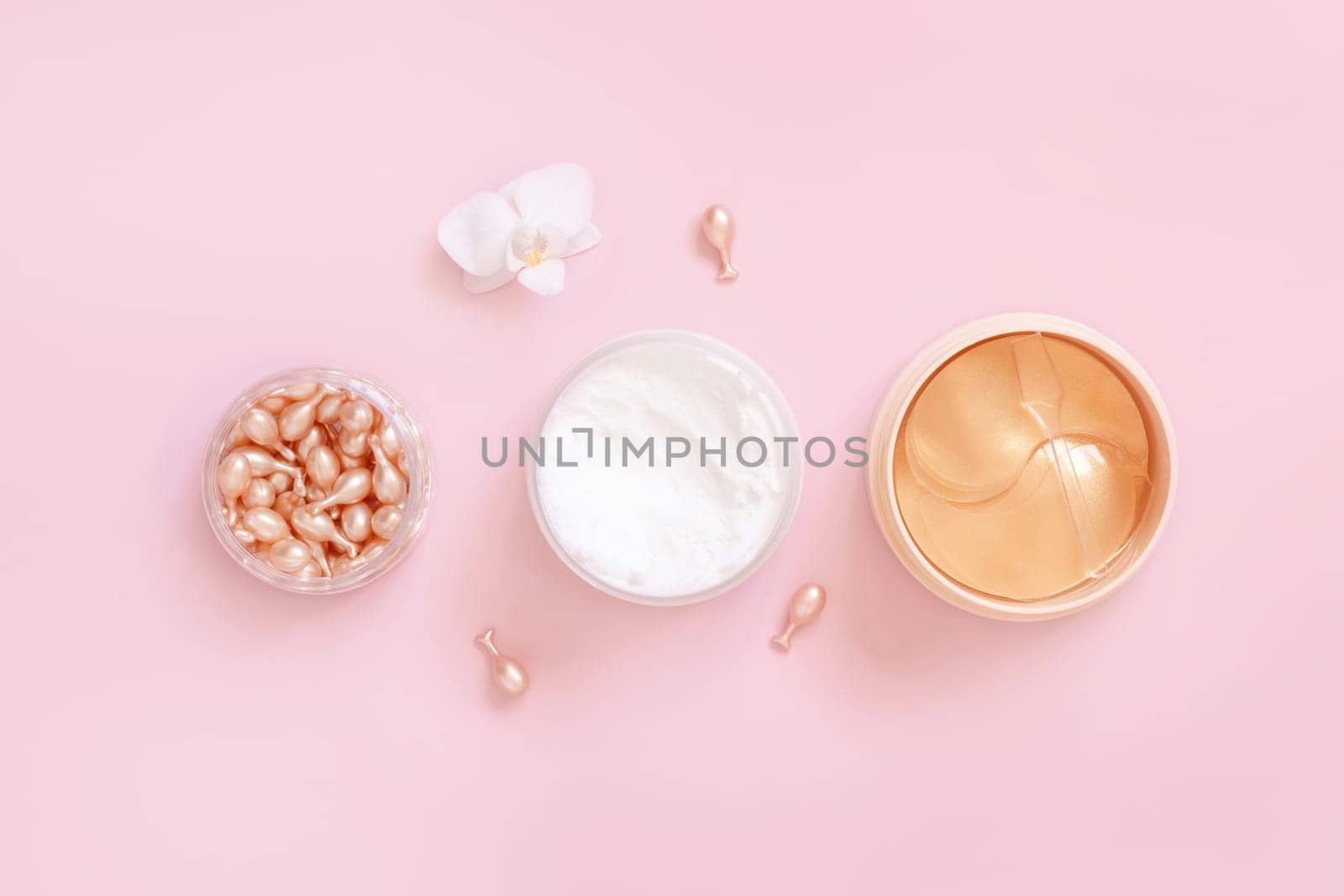 Face serum capsules, cream and eye patches on light pink near orchid flower top view. Skincare cosmetics. Facial Beauty treatments. Pastel minimal composition..
