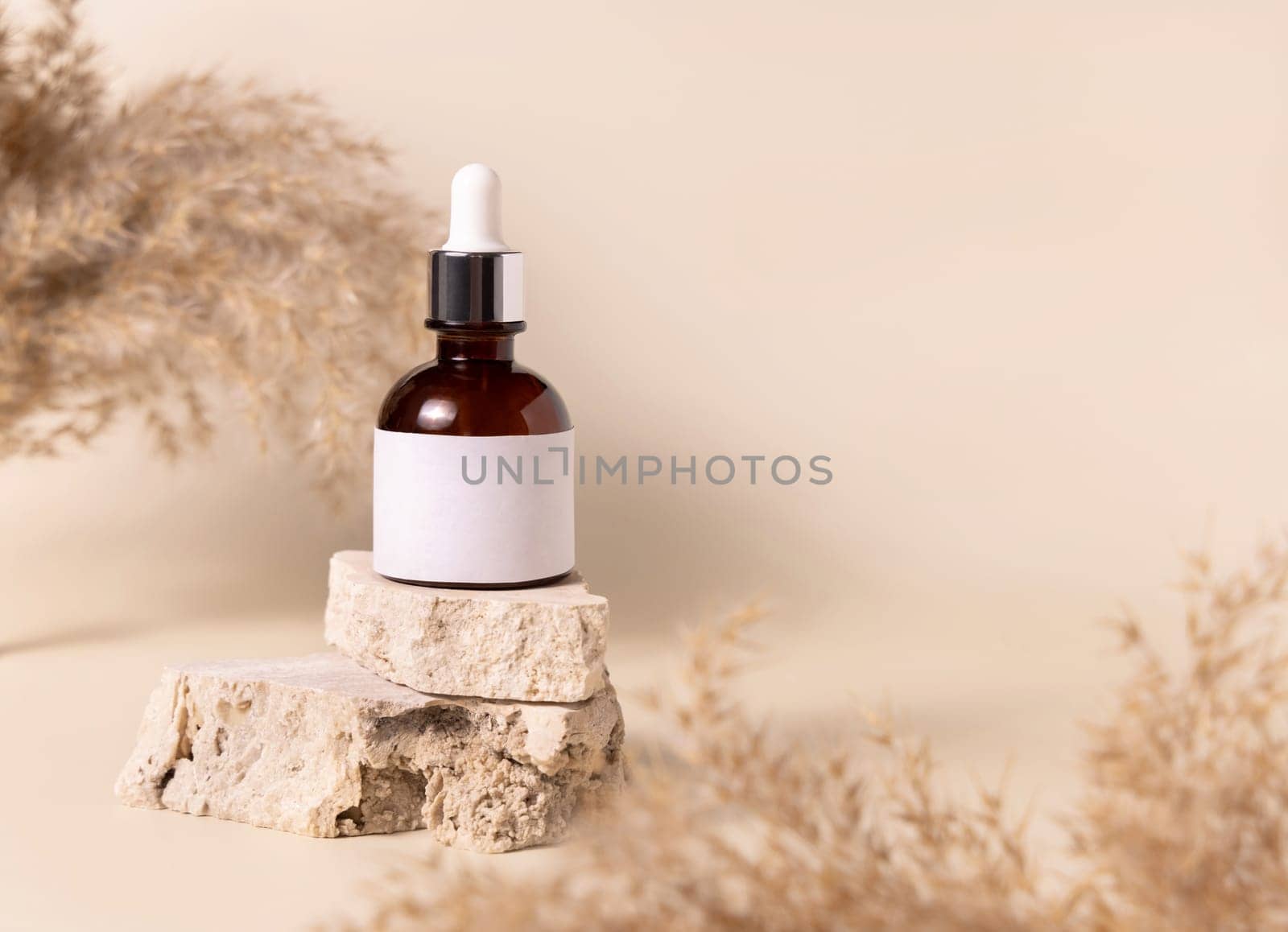 Brown dropper bottle with blank label on travertine stone near dry pampas grass on light yellow, close up, mockup. Skincare beauty product. Natural cosmetics. Boho composition..
