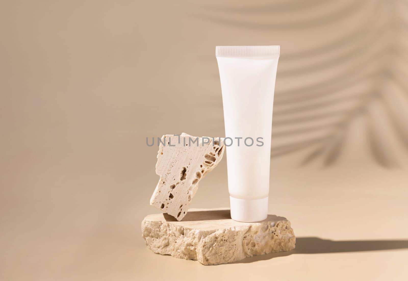 White plastic cream tube on travertine stone on light yellow, Palm leaf hard shadows, close up, mockup. Skincare beauty product. Natural cosmetics. Boho composition..