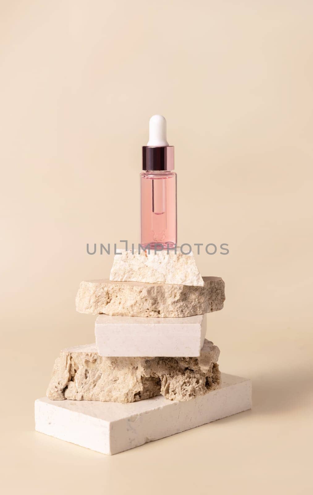 Pink glass cosmetic dropper bottle on beige travertine stones on light yellow close up, mockup. Skincare natural beauty product. Minimal eco friendly composition.