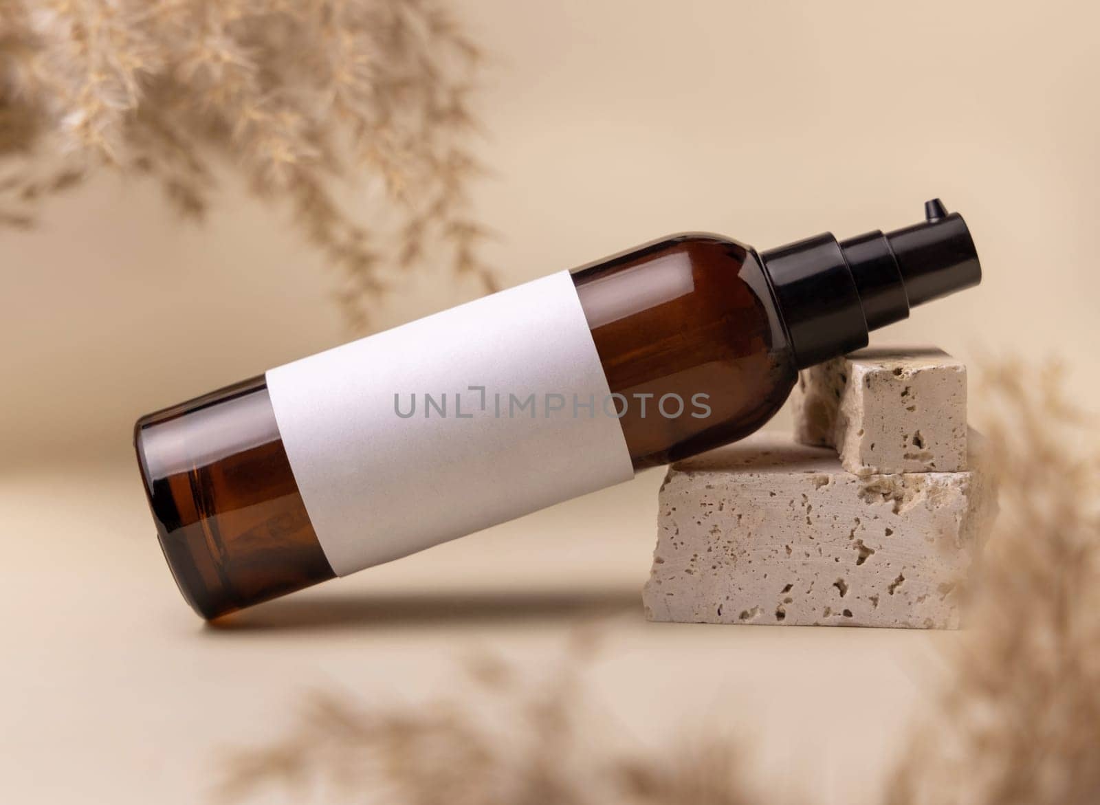 Brown glass cosmetic bottle with blank label on stones near pampas grass on light yellow, close up, mockup. Skincare beauty product. Natural eco friendly cosmetics